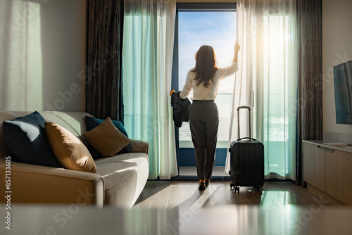 Confident businesswoman wearing white shirt and holding suit with suitcase stands in a hotel room while looking at the view from window Beautiful sunset. Woman enjoying her holiday in a luxury hotel.