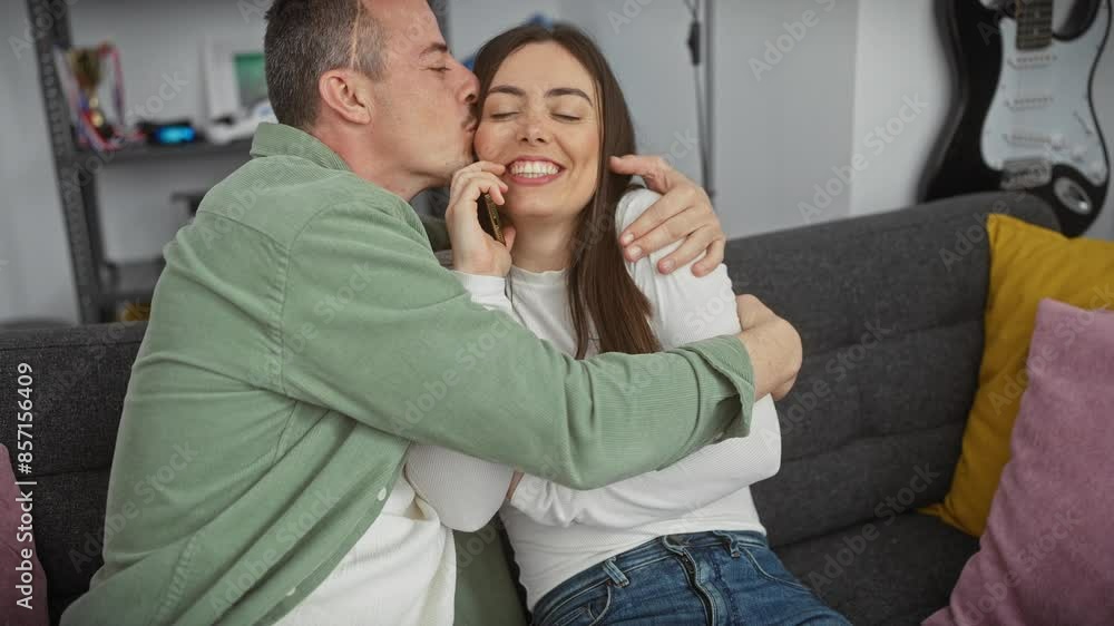 A loving couple enjoys a cozy moment at home, with the man kissing the smiling woman as she talks on the phone A loving couple enjoys a cozy moment at home, with the man kissing the smiling woman as she talks on the phone