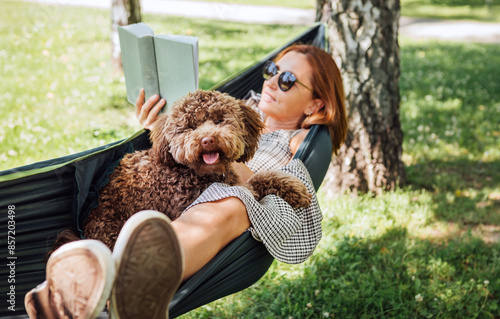 Woman reading book relaxing in hammock with her fluffy brown Maltipoo dog on sunny day. Both looking content and happy. This outdoor scene captures joy of bonding with pets and enjoying togetherness