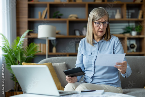 Senior woman looking worried while managing finances at home, using a laptop and holding documents. Concept of financial stress, budgeting, and retirement planning.