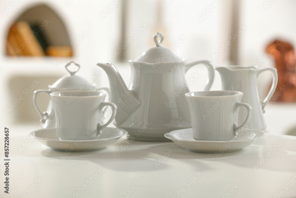 Set of different dishes and cups and saucers and coffee pot on white table top with blurred white kitchen interior Set of different dishes and cups and saucers and coffee pot on white table top with blurred white kitchen interior