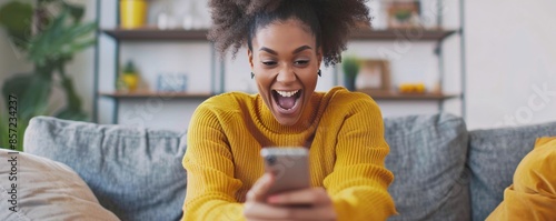 Young woman sitting on the couch at home, looking pleasantly surprised and happy while holding a smartphone photo