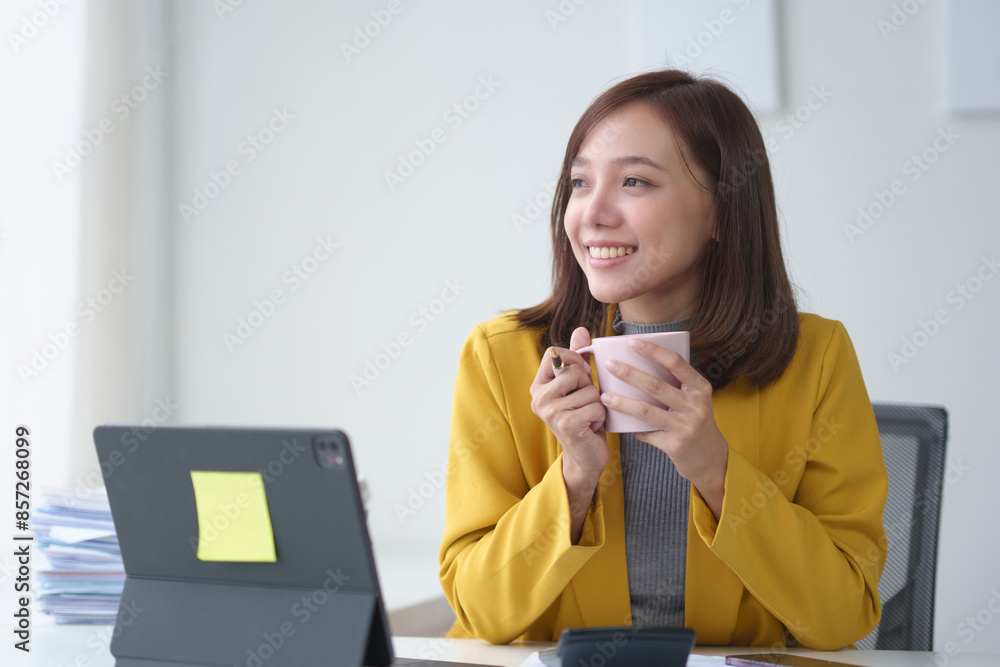 Businesswoman in yellow blazer smiling while holding coffee cup, sitting at desk with laptop in a bright office setting. Businesswoman in yellow blazer smiling while holding coffee cup, sitting at desk with laptop in a bright office setting.