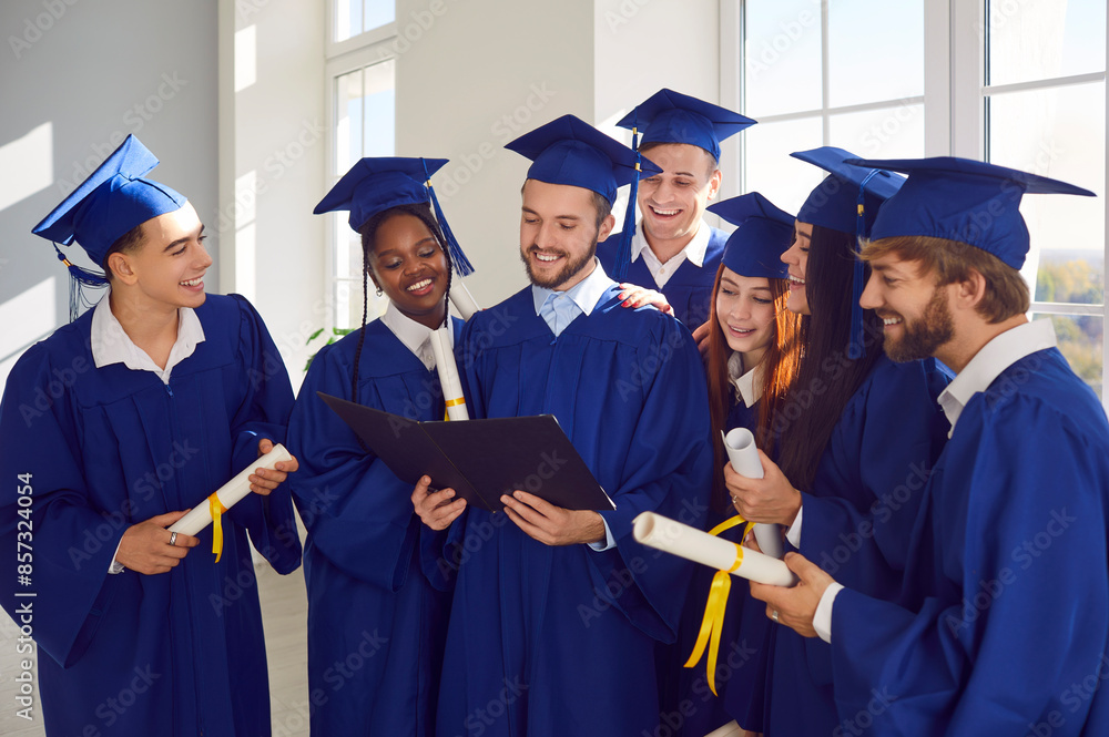 Happy smiling diverse multiethnic graduate students in blue graduation robes celebrate graduation and looking to diploma of their classmate standing indoors. Education and graduation concept. Happy smiling diverse multiethnic graduate students in blue graduation robes celebrate graduation and looking to diploma of their classmate standing indoors. Education and graduation concept.