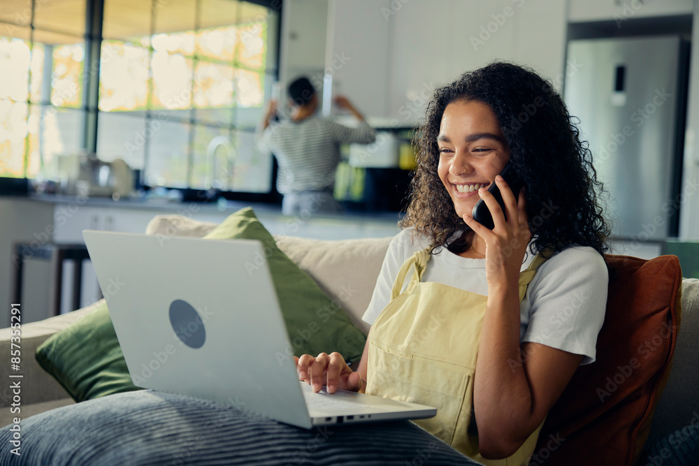 Smiling woman sitting on couch with laptop on thigh, talking on cell phone Smiling woman sitting on couch with laptop on thigh, talking on cell phone