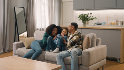 Happy excited African American family relocation woman female mom little boy child son kid father male man moving couch furniture fall jump on sofa celebrating buying new home modern apartment indoors
