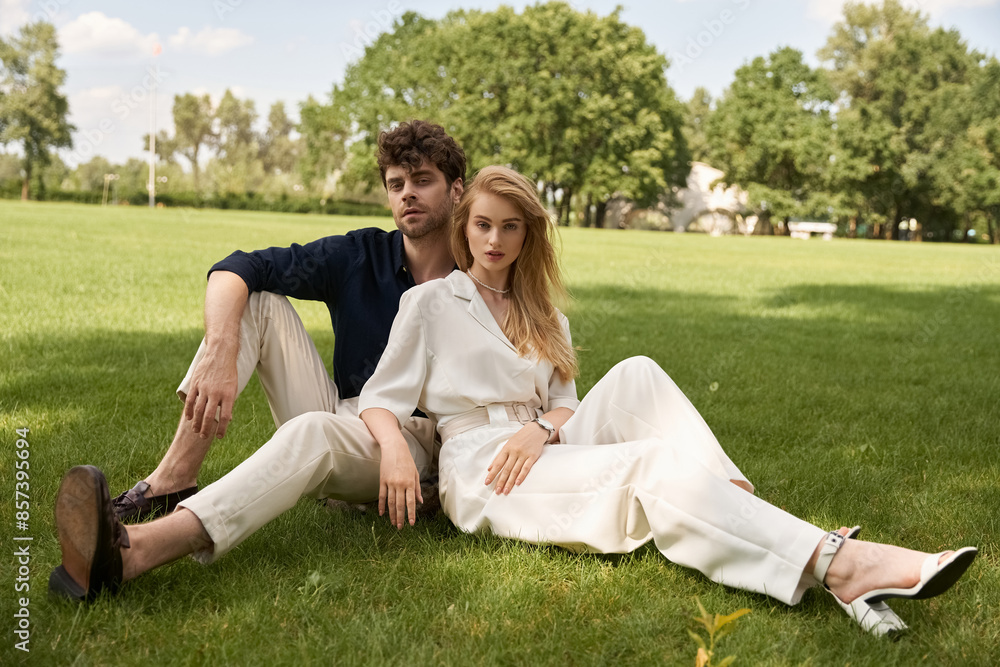 Elegant couple in refined attire enjoying a leisurely day sitting on grass in a lush park.