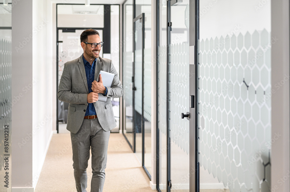 Portrait of handsome project manager with laptop and glasses smiling and walking in office corridor Portrait of handsome project manager with laptop and glasses smiling and walking in office corridor