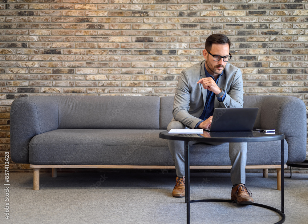Full length of serious young manager reading e-mails over laptop while sitting on sofa in office Full length of serious young manager reading e-mails over laptop while sitting on sofa in office