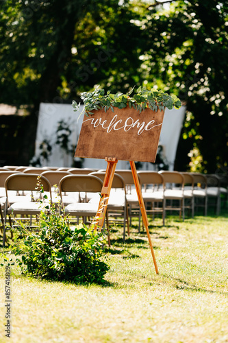 Beautiful outdoor wedding setting with a welcome sign on an easel, surrounded by chairs and greenery.