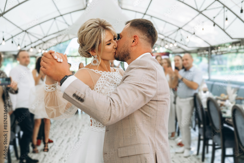 Bride and groom dancing the first wedding dance and kisses against the background of guests in an evening restaurant Bride and groom dancing the first wedding dance and kisses against the background of guests in an evening restaurant