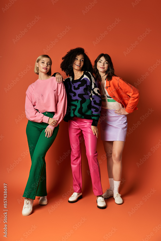 Fototapeta premium Three young beautiful multicultural women in stylish vibrant clothes stand united on an orange background.