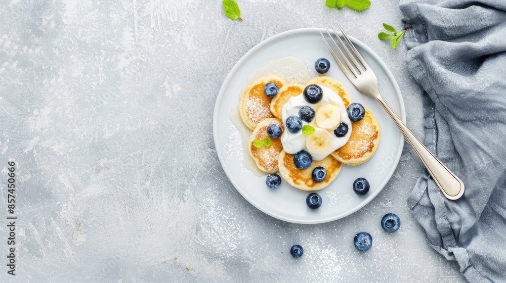 A plate of golden brown pancakes topped with whipped cream and blueberries, sitting on a white background with a fork and knife nearby A plate of golden brown pancakes topped with whipped cream and blueberries, sitting on a white background with a fork and knife nearby