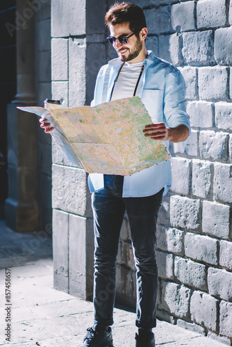 Positive bearded casual dressed tourist in stylish sunglasses looking at travel map and searching location of showplaces while walkig in urban setting of architectural downtown with coffee to go photo