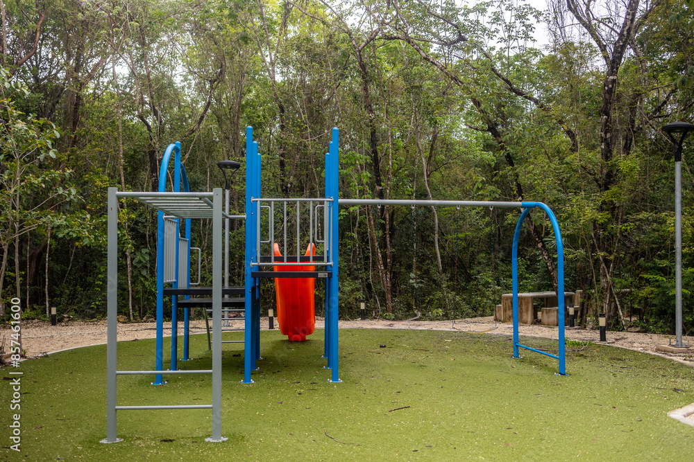 Fototapeta premium Blue and red kids playground with slide with fake green grass surrounded by tropical jungle on a cloud day