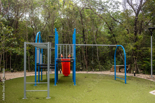 Blue and red kids playground with slide with fake green grass surrounded by tropical jungle on a cloud day 