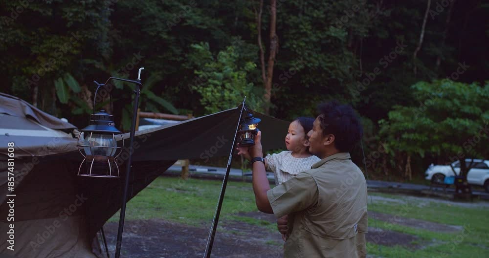 A father holding his child Enjoying Camping Trip in Forest with illuminated by lanterns at dusk, The scene captures a serene and bonding moment in nature. A father holding his child Enjoying Camping Trip in Forest with illuminated by lanterns at dusk, The scene captures a serene and bonding moment in nature.