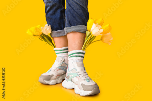 Legs of beautiful young woman in sneakers with tulips flowers on yellow background #857531210