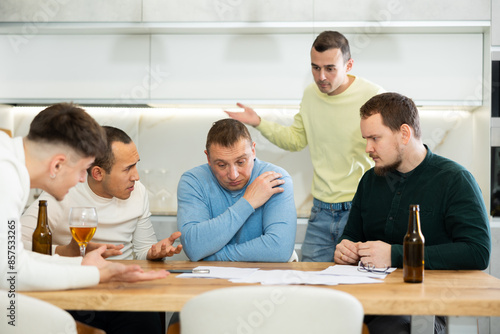Upset worried man sitting at table in home kitchen with papers in hands, reading unexpected medical results with group of male friends solacing and comforting him .... © JackF