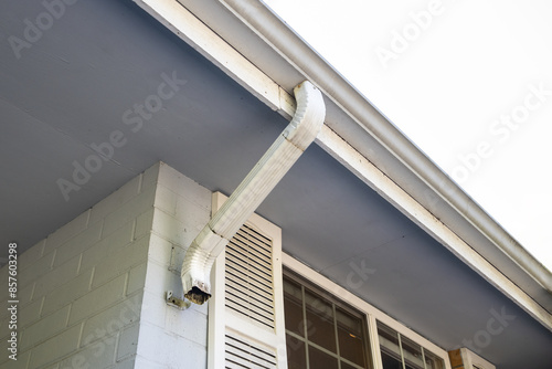 Rectangular white metal rainwater downspout detached from mounting bracket and sections pulled apart, house with painted brick wall, window and shutter
