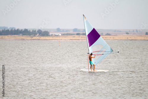 Deportista practicando windsurf en el mar, captado en plena acción bajo un cielo despejado. La colorida vela contrasta con las olas, transmitiendo energía y emoción.