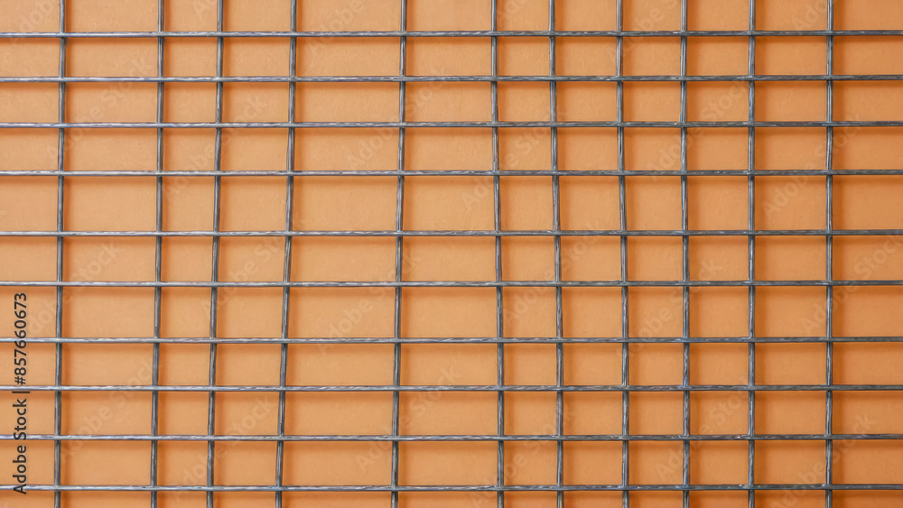 Lattice made of metal thin wire sits against orange background. Grid features both square and rectangular cells, offering unique geometric pattern. Top view of grating. Lattice made of metal thin wire sits against orange background. Grid features both square and rectangular cells, offering unique geometric pattern. Top view of grating.