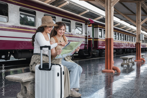 Two young Asian women sitting on a bench at a train station, holding a map and pointing, with a suitcase beside them, planning their travel route with excitement