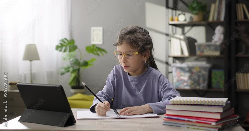 Cute little girl study online watching webinar educational tutorial using app on digital tablet, doing homework, virtual lesson class, making notes in a copybook sitting at the desk Cute little girl study online watching webinar educational tutorial using app on digital tablet, doing homework, virtual lesson class, making notes in a copybook sitting at the desk