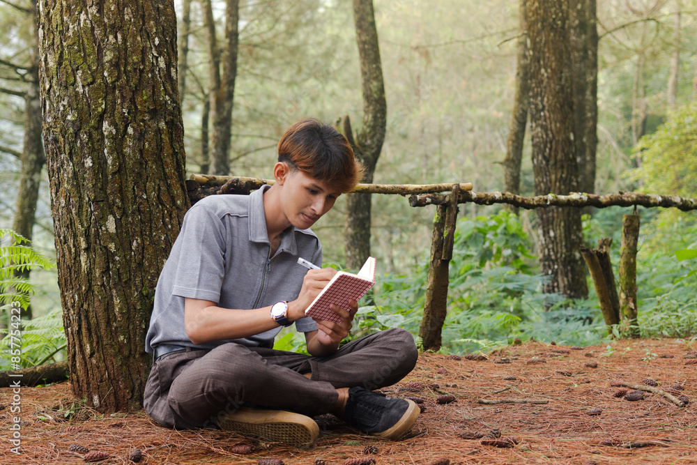 Fototapeta premium Portrait of confident Asian man writing on notebook while thinking and looking away in the nature forest. Peaceful and relaxing healthcare wellness