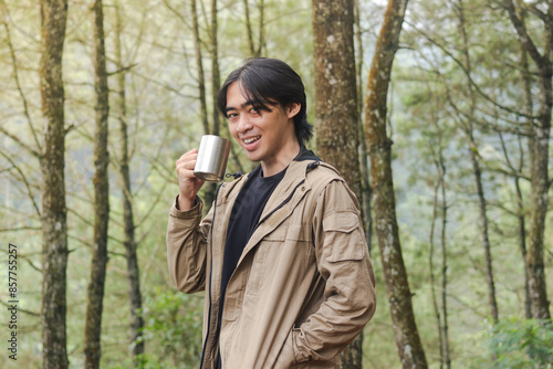 Portrait of excited Asian hiker man drinking water from stainless steel cup while trekking in nature forest