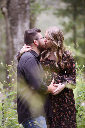 A pregnant couple kissing in a forest, with the man holding the photo