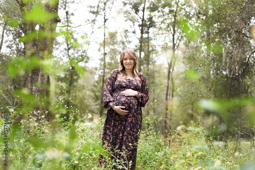 Pregnant woman in a floral dress standing in a forest, holding h photo
