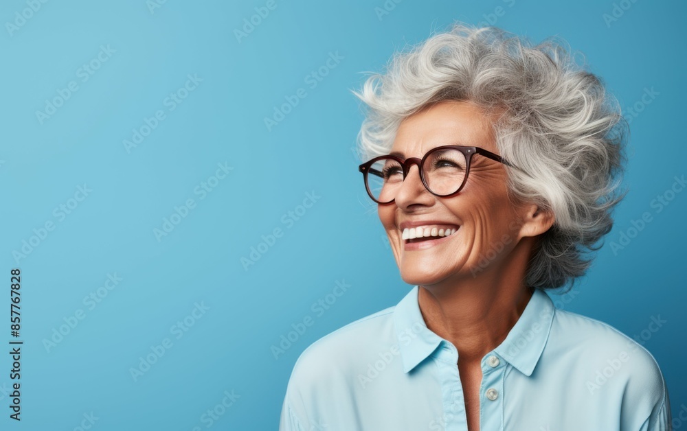 A woman with glasses is smiling and looking at the camera. She is wearing a blue shirt and has a blue background A woman with glasses is smiling and looking at the camera. She is wearing a blue shirt and has a blue background