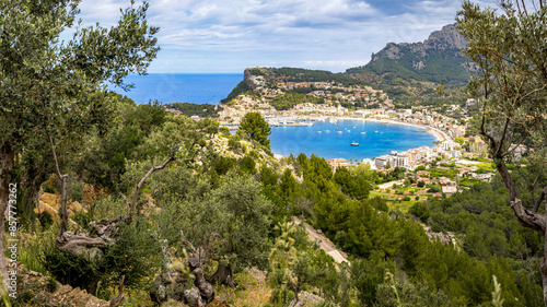 Panoramic view framed by olive trees over the bay of Port de Sóller from the GR-221 hiking trail with Puig de Bàlitx mountain in the background on an overcast day, ideal for nature tourism in Mallorca