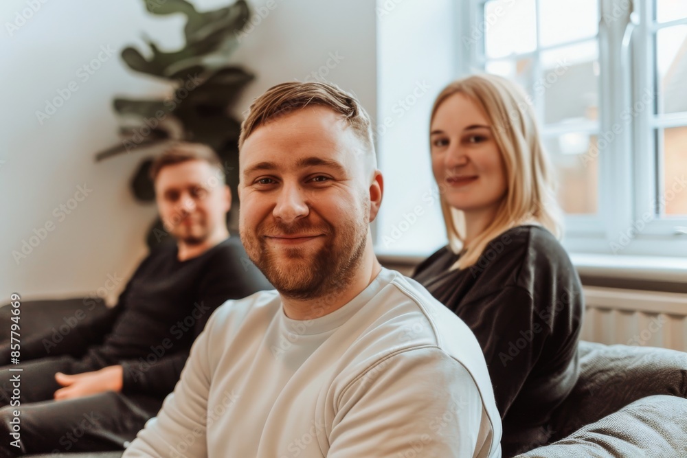 Portrait of smiling man sitting on sofa and looking at camera with his colleagues in background Portrait of smiling man sitting on sofa and looking at camera with his colleagues in background