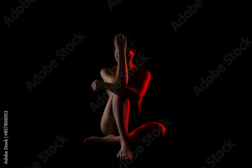 The image shows a woman sitting in a dark studio with red light illuminating her body. She is facing the camera with her hands clasped together near her chest, and her head is slightly tilted down