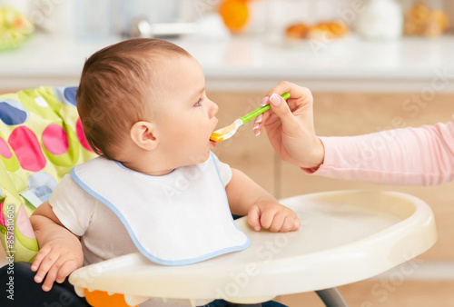 Bilde på lerret A mother is feeding her baby boy who is seated in a high chair in the kitchen