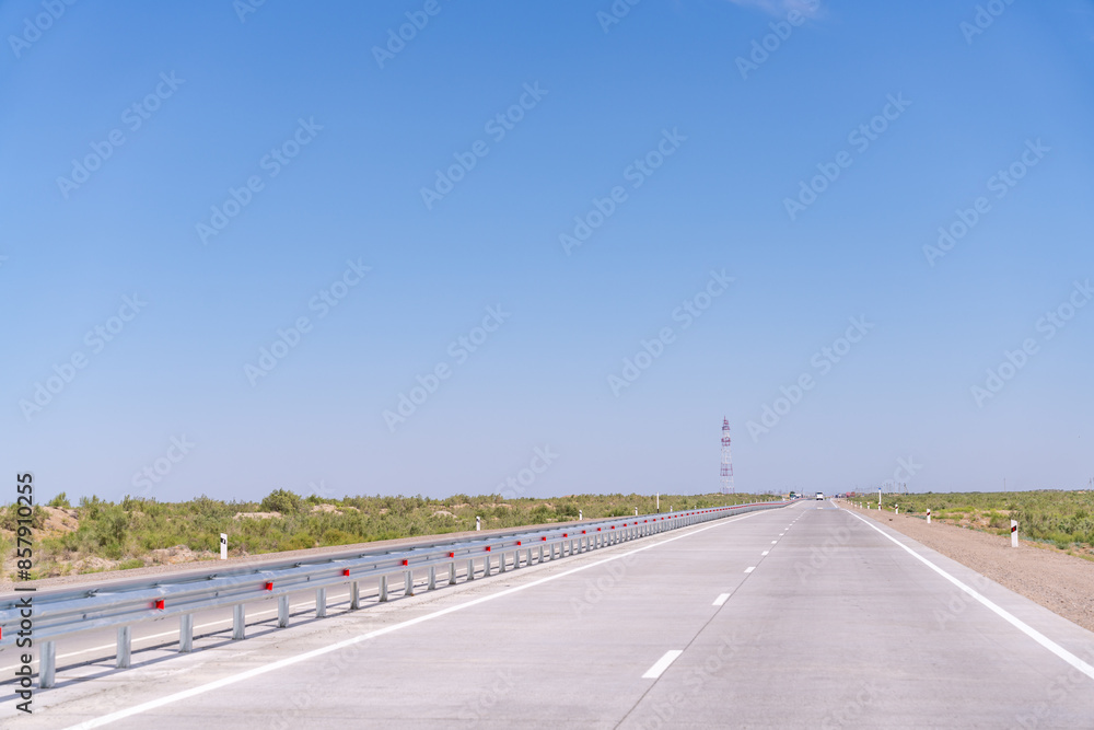 A long, empty road with a blue sky above. The road is lined with a metal fence A long, empty road with a blue sky above. The road is lined with a metal fence