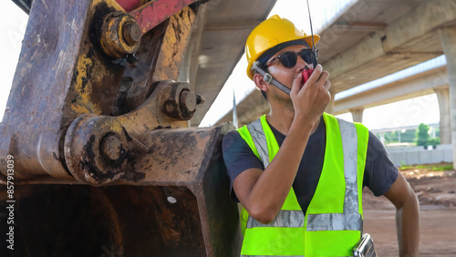 Asian male engineer standing next to an excavator Use the radio to check and order work at Construction size: highway bridge photo