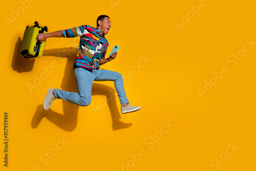 Full body photo of attractive young man suitcase run hurry hold device dressed stylish colorful clothes isolated on yellow color background