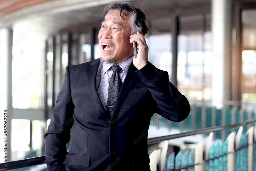 Stressed senior Asian businessman in suit having panic attack while talking on a mobile phone. Elderly business owner is shocked by bad news. Mature boss getting mad while talking on the phone. Stressed senior Asian businessman in suit having panic attack while talking on a mobile phone. Elderly business owner is shocked by bad news. Mature boss getting mad while talking on the phone.