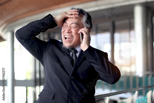 Stressed senior Asian businessman in suit having panic attack while talking on a mobile phone. Elderly business owner is shocked by bad news. Mature boss getting mad while talking on the phone.