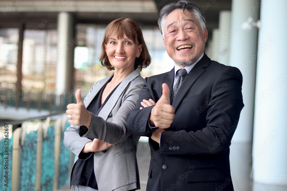 Happy smiling senior Asian businessman in suit with Caucasian businesswoman giving thumbs up to camera at office. Elderly successful male and female boss at workplace. Portrait mature businesspeople. Happy smiling senior Asian businessman in suit with Caucasian businesswoman giving thumbs up to camera at office. Elderly successful male and female boss at workplace. Portrait mature businesspeople.