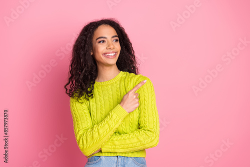 Portrait of beautiful curly haired brunette beginner saleswoman in green knitted pullover pointing finger promo isolated on pink color background