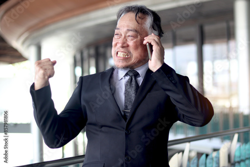 Happy senior Asian businessman in suit showing clenched fist winner gesture. Surprised elderly business does a winning closed while talks on mobile phone. Mature boss gesturing yes with clenched fist.