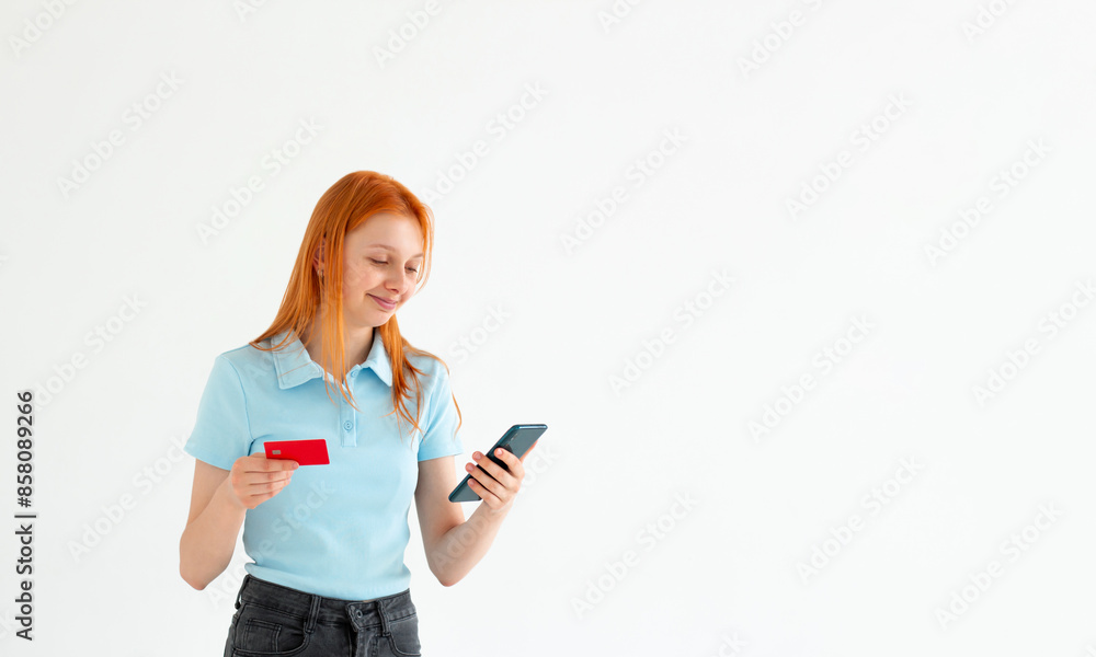 Cheerful young girl showing plastic credit card while using mobile phone isolated over white background, wearing blue t-shirt Cheerful young girl showing plastic credit card while using mobile phone isolated over white background, wearing blue t-shirt