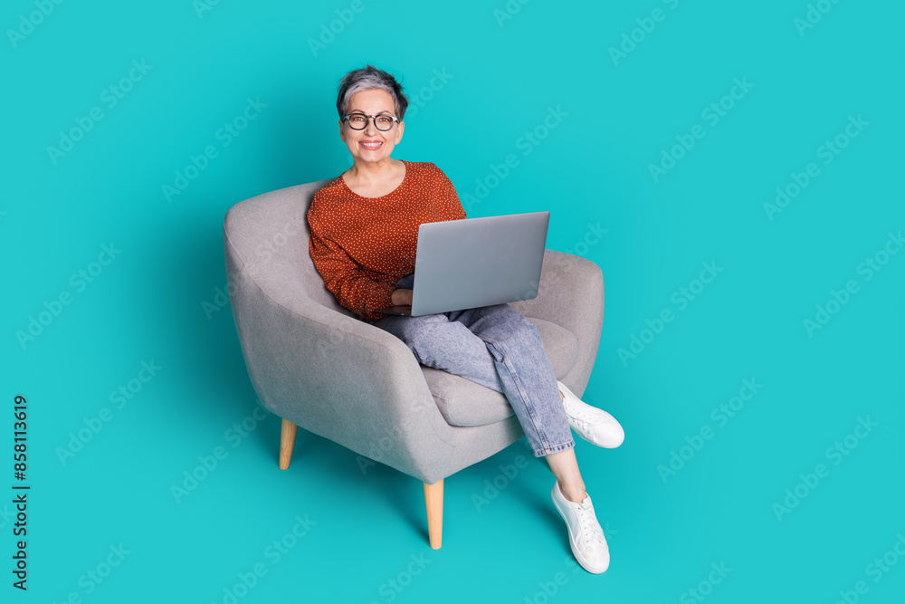 Full length photo of lovely pensioner lady sit armchair work netbook dressed stylish brown garment isolated on cyan color background Full length photo of lovely pensioner lady sit armchair work netbook dressed stylish brown garment isolated on cyan color background