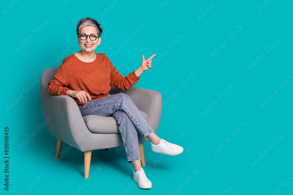 Full length photo of lovely pensioner lady sit armchair point empty space dressed stylish brown garment isolated on cyan color background Full length photo of lovely pensioner lady sit armchair point empty space dressed stylish brown garment isolated on cyan color background