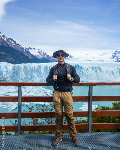A young man with a mustache wearing a hat, sunglasses, and trekking clothes poses in front of the Perito Moreno glacier, Argentina.