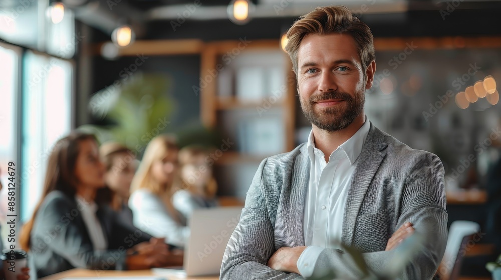 A man with a beard is confidently leading a team meeting in an office, standing with arms crossed while colleagues are discussing in a well-lit, modern workspace.